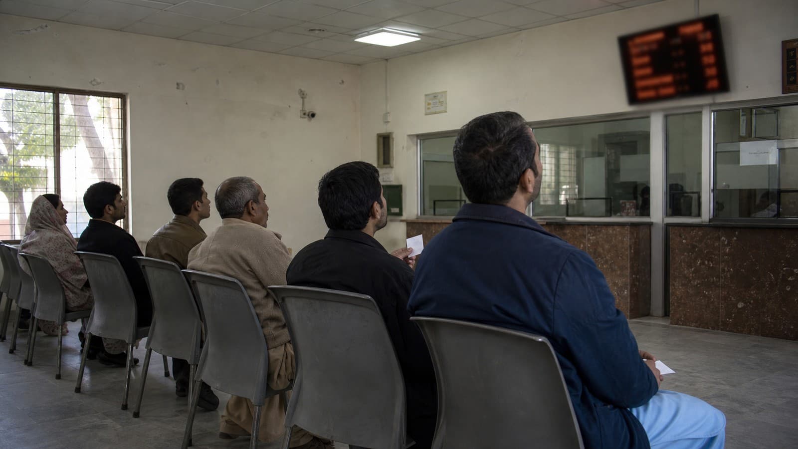 Customers seated in a waiting hall holding numbered tokens, looking towards a counter display board