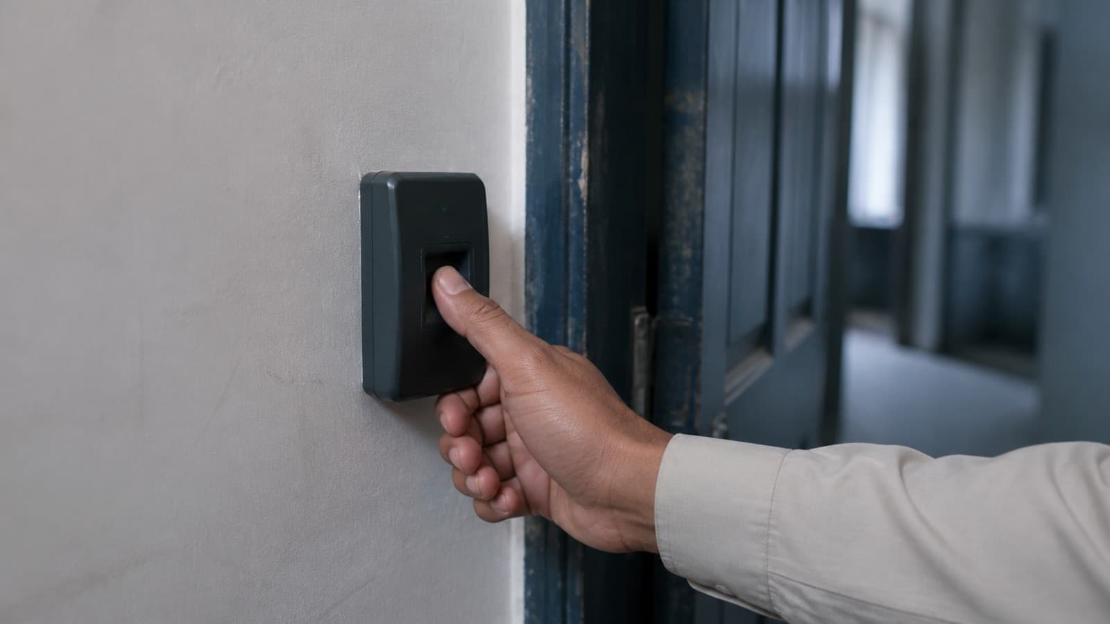 A hand pressing a thumb onto a fingerprint access control reader mounted beside an office door